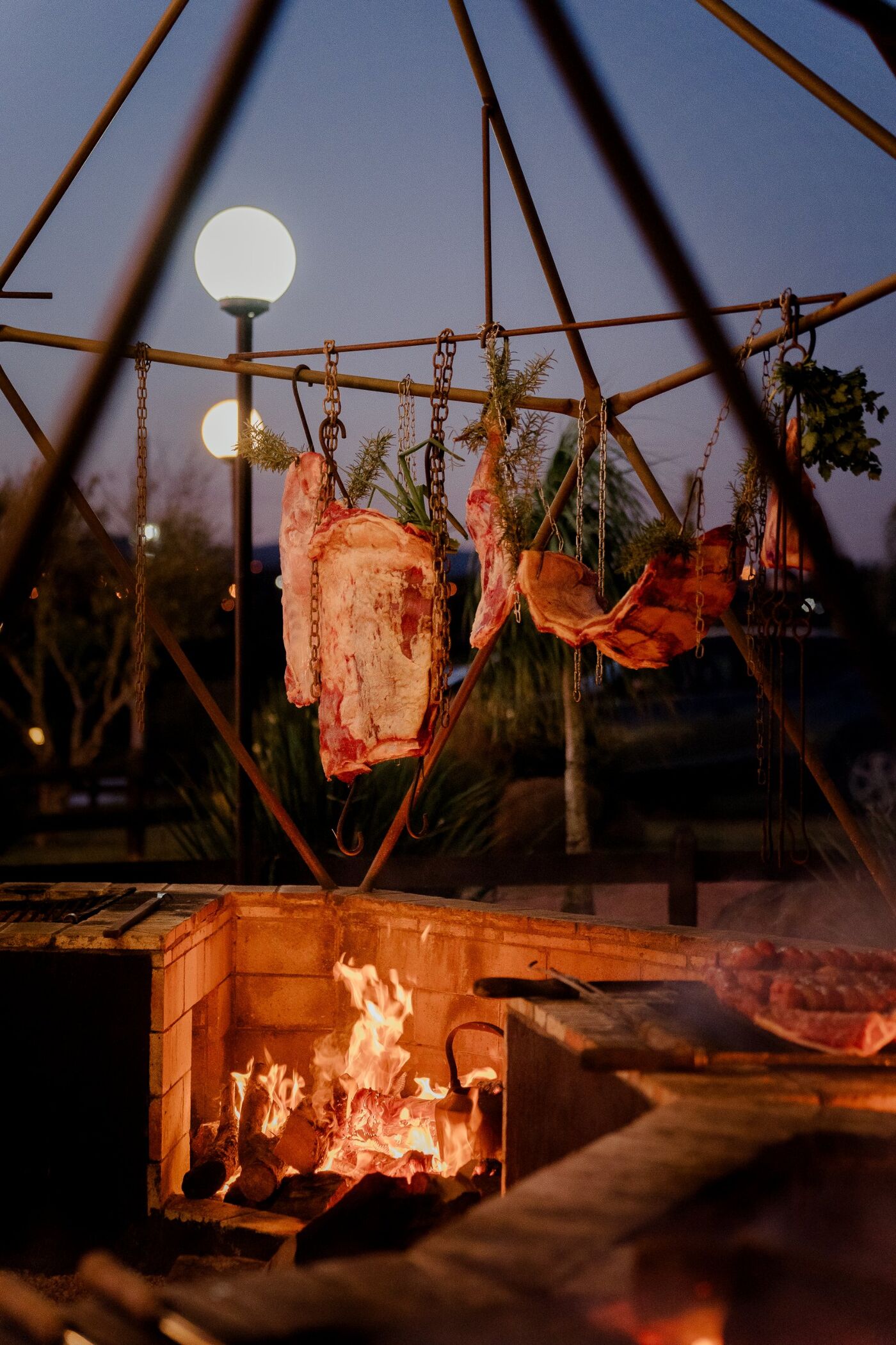 Cortes pendurados na estrutura de parrilla sobre o fogo de chão, ao anoitecer